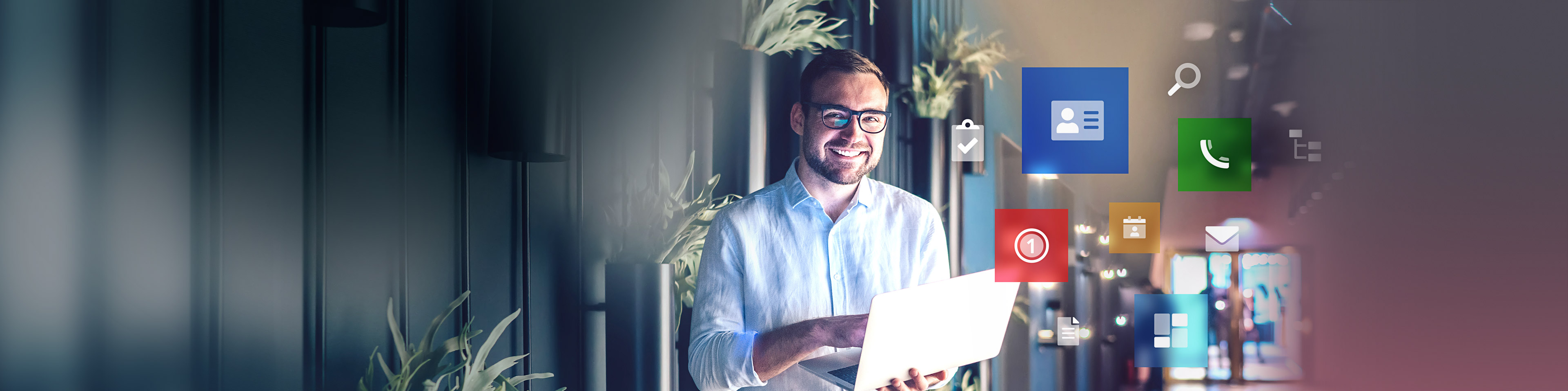 Lächelnder Mann steht mit aufgeklapptem Laptop im modernen Flur eines Bürokomplexes. Rechts neben ihm schweben verschiedene Produkticons in unterschiedlichen Farben und Größen.