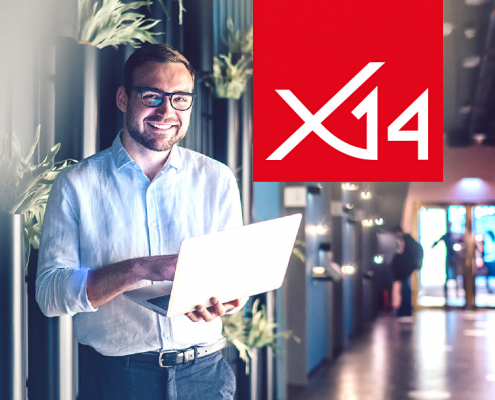 Lächelnder Mann steht mit aufgeklapptem Laptop im modernen Flur eines Bürokomplexes. Rechts oben das x14-Logo in weiß auf einem roten Kasten.